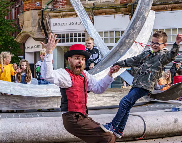 A bearded man in colourful hat and waistcoat leaning back supporting a young boy standing on his knees