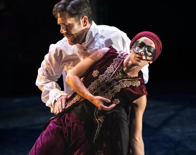 Two dance performers, one dressed in white Edwardian shirt, the other masked and wearing an Arabian embroidered sleeveless jacket