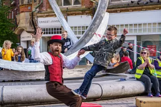 A bearded man in colourful hat and waistcoat leaning back supporting a young boy standing on his knees